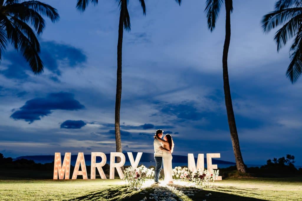 William and Roshni lit up while the sun has set and the Marry Me marquee letters are illuminating behind them