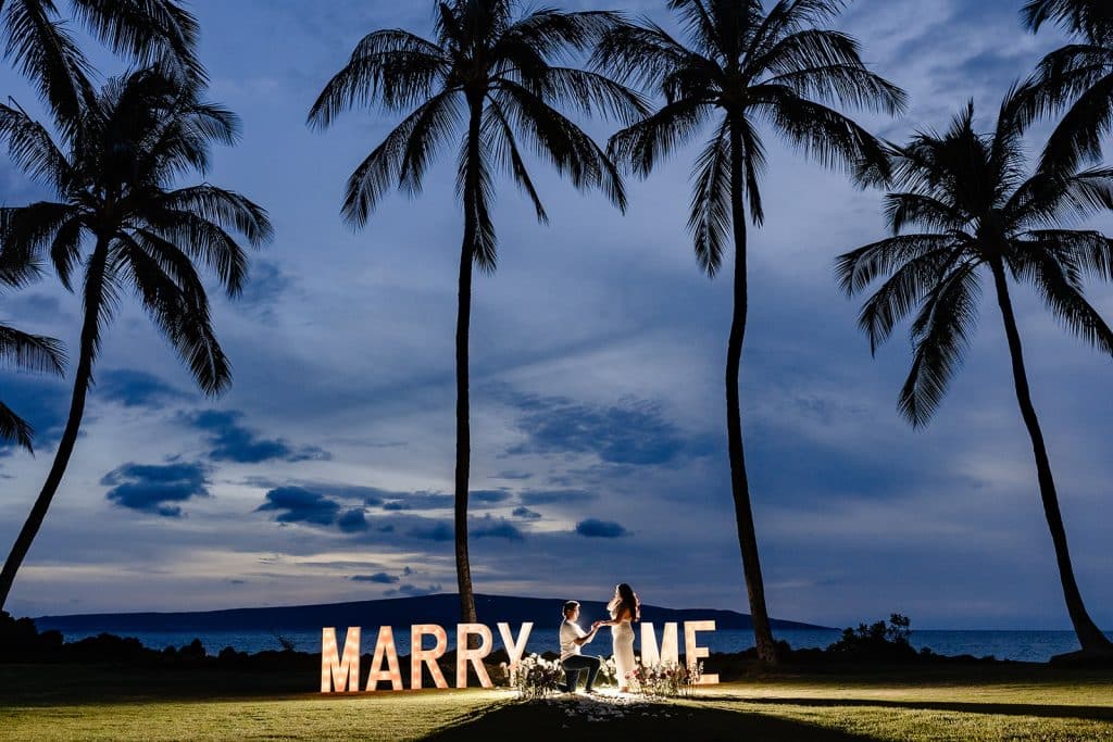 William on one knee and recreating his proposal to Roshni while the sun has gone down and the Marry Me letters are lit up