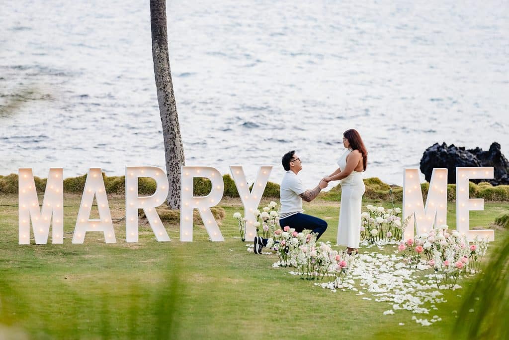 William on one knee proposing to Roshni in the middle of the Marry Me marquee letters and surrounded by flowers and flower petals