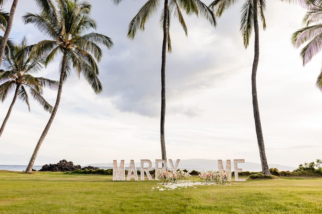 A view of the Marry Me marquee letters little up with white flower petals and white and pink flowers lining the pathway. Palm trees and the ocean are in the background as the sun starts to set.