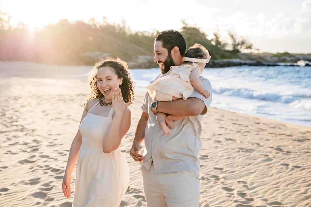 Andy holding their baby and smiling at Tanya as she poses for the camera with her hand up to show off her new ring