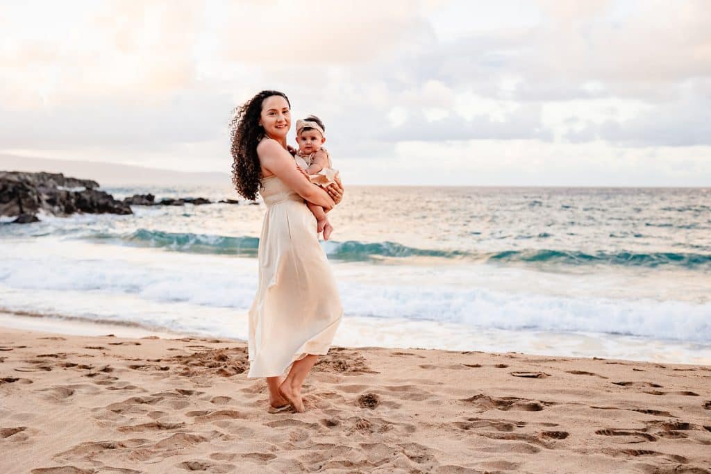 Tanya walking through the sand towards the water while holding her baby girl and they're both smiling at the camera