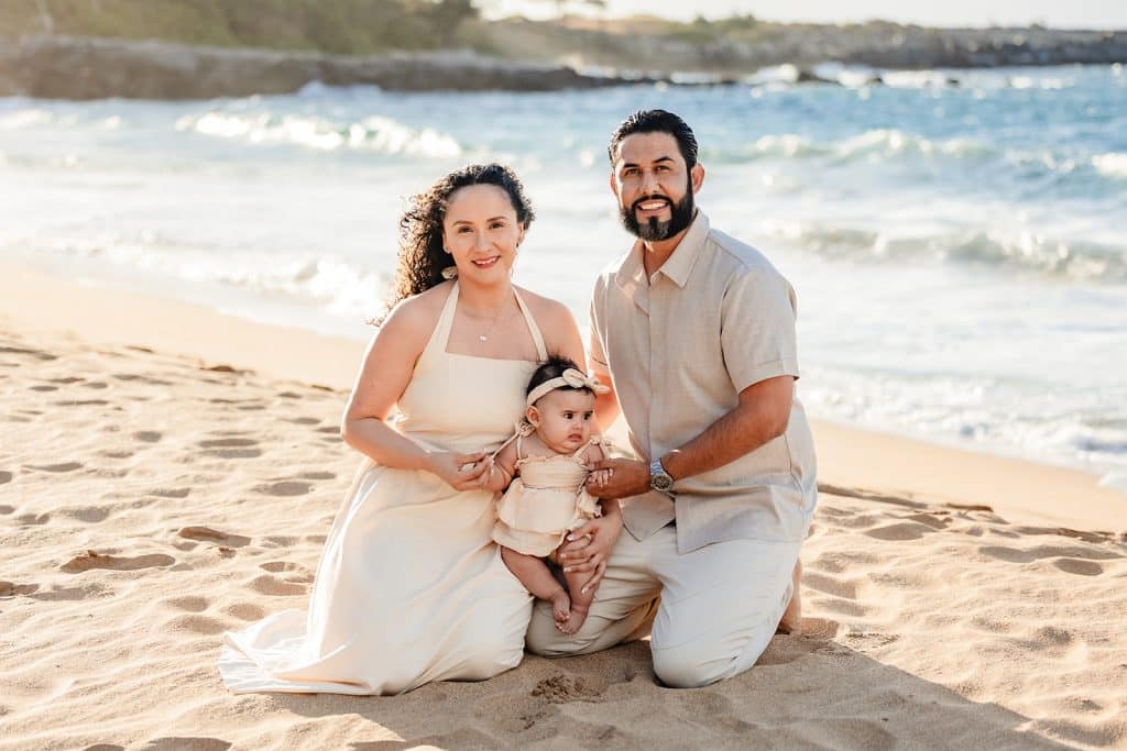 Andy and Tanya kneeling in the sand on the beach at sunset with their baby girl sitting on their legs in between them.