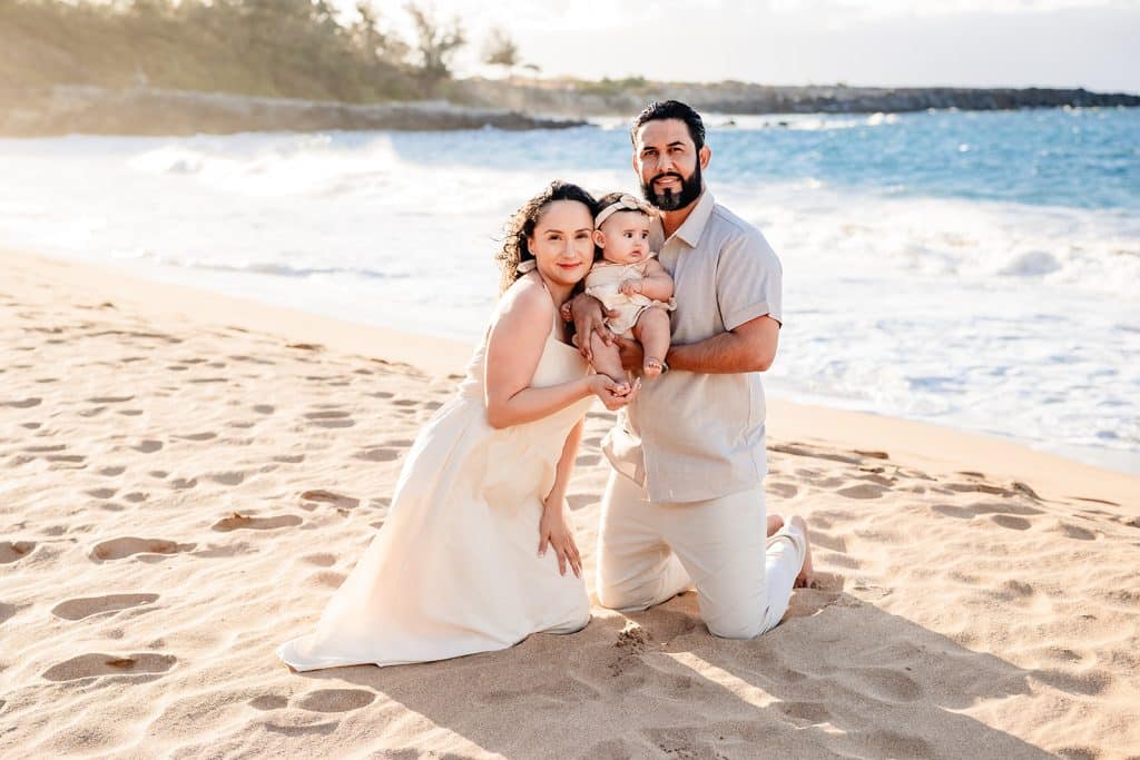 Andy and Tanya kneeling in the sand on the beach at sunset with their baby girl in between them