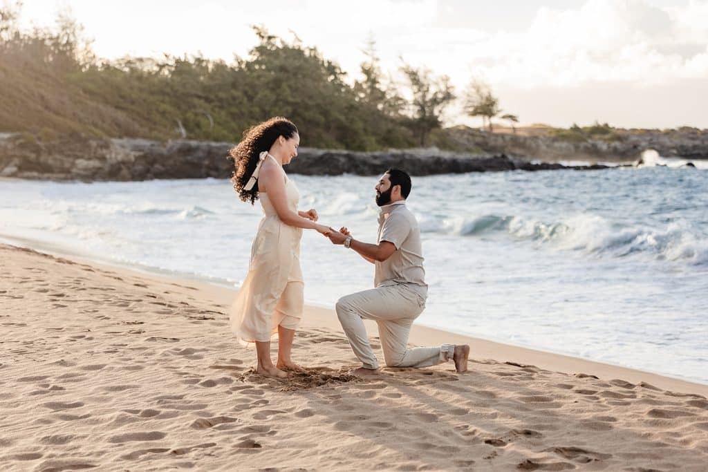 Andy down on one knee in the sand and holding Tanya's hand as she looks down at him smiling.