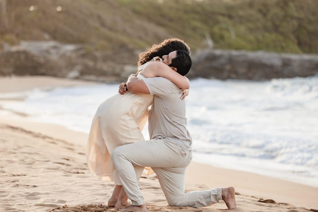 A closeup of Tanya leaning down and hugging Andy in the sand while he is still down on one knee.