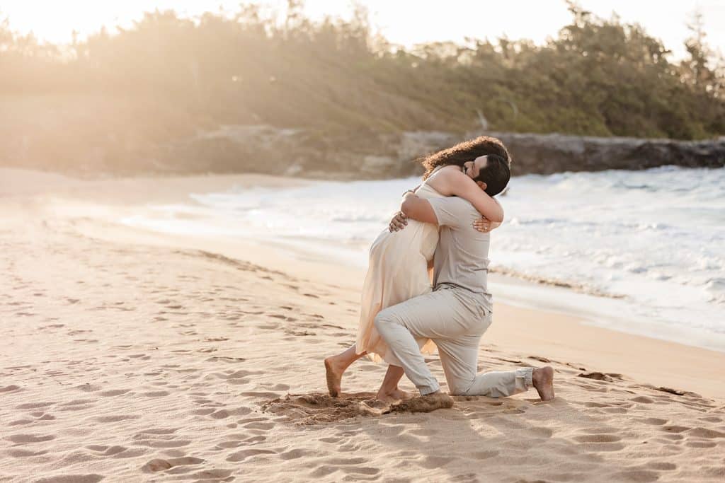 Tanya leaning down and hugging Andy in the sand while he is still down on one knee.