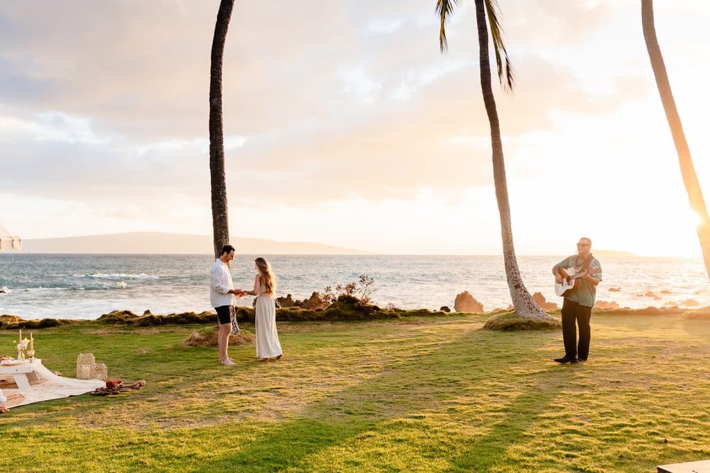 A wide angle shot of Patrick and Rachel admiring the ring while the musician plays guitar softly. The sun is setting over the ocean in the background.