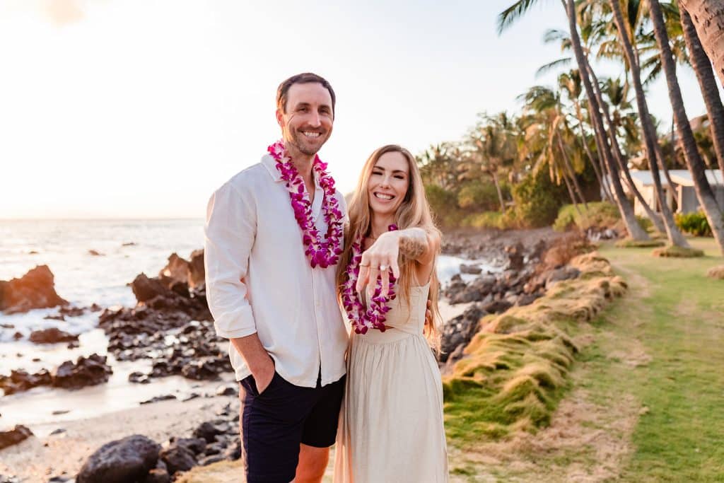 Patrick and Rachel smiling at the camera while Rachel holds out her hand to show off her new ring. The ocean and palm trees are behind them.