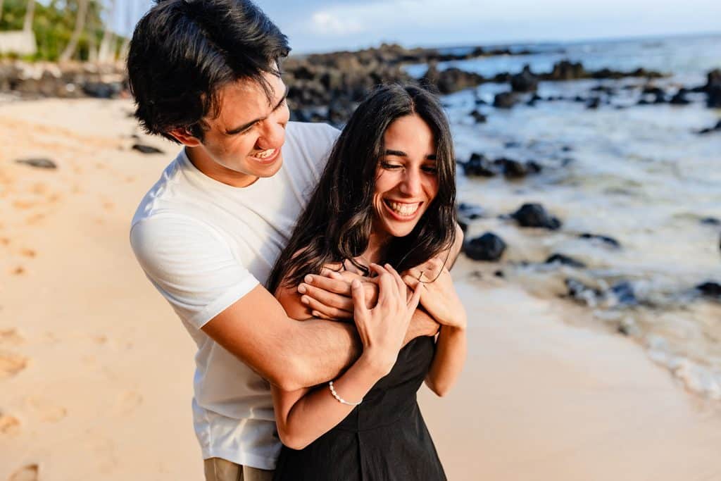 Hector hugging Alejandra from behind as she grabs his arms around her chest. They're both laughing.