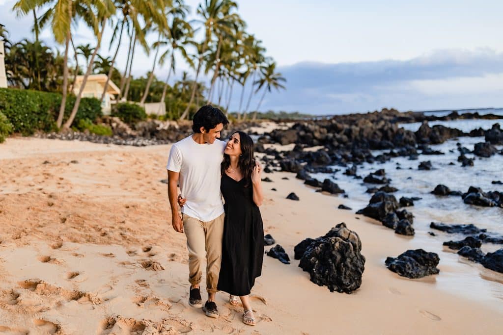 Hector and Alejandra walking along the shore at sunset. His arm is over her shoulder and they're looking at each other and smiling.