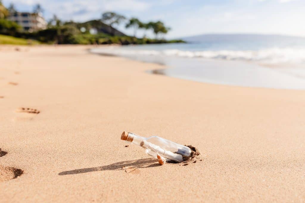 A close up photo of a glass message in a bottle sticking out of the sand as the waves crash into the shore in the background.