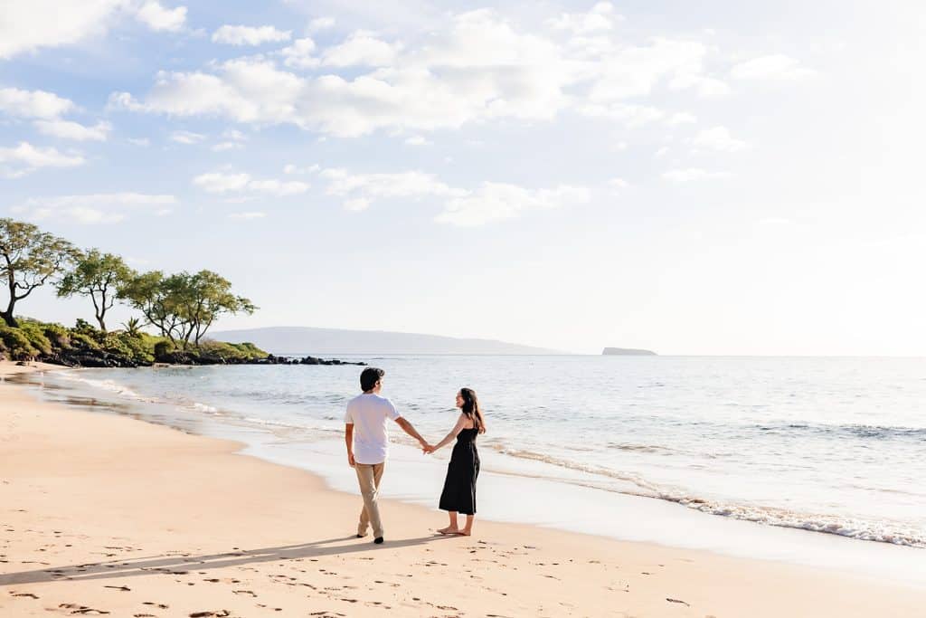 Hector and Alejandra walking along the shore holding hands. They're walking farther from the camera as they laugh and smile at each other.