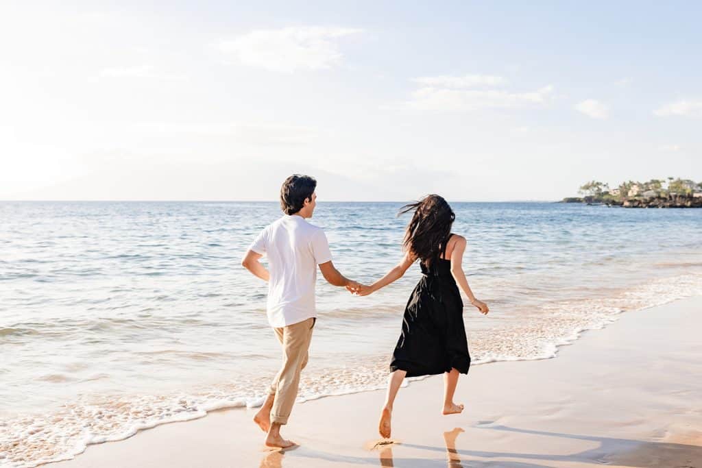 The back of Alejandra and Hector running along the shoreline and holding hands while the sun is setting.