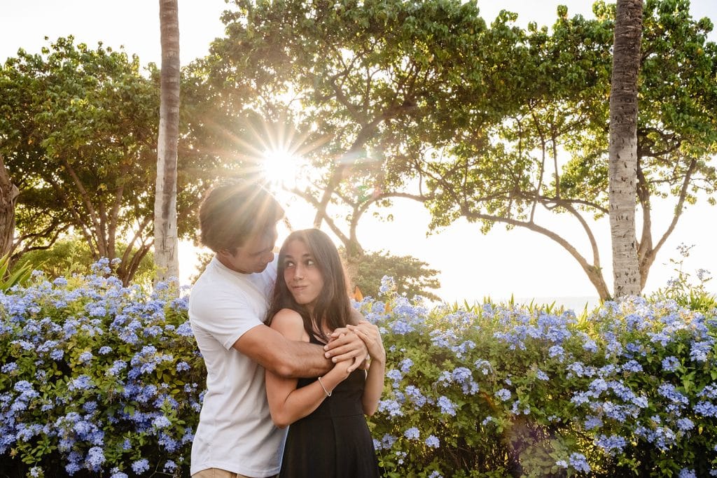 Hector hugging Alejandra from behind as she looks up at him and smiles. The sun is setting behind them and there are bushes of periwinkle flowers all around them.