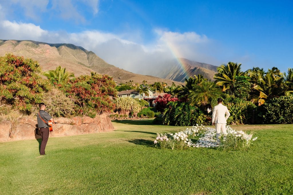 A view of the back of Pruthvi and the musician. There are mountains and a rainbow overhead where Tithi will walk in.