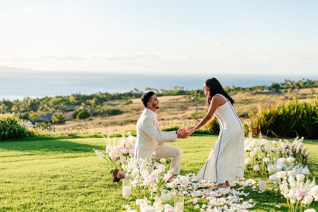 Pruthvi down on one knee among the flowers and candle as the ocean is in view behind them. Tithi is holding his hands and smiling.