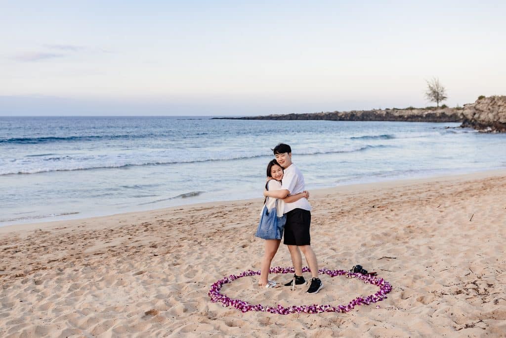 Shunshun and Aaron hugging and looking at the camera surrounded by orchids in the sand.
