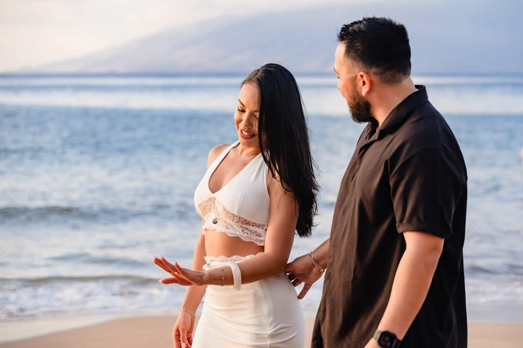 Brandon and Alexis are walking the shoreline while she looks down at her hand with a big smile. Brandon is looking at her and smiling.