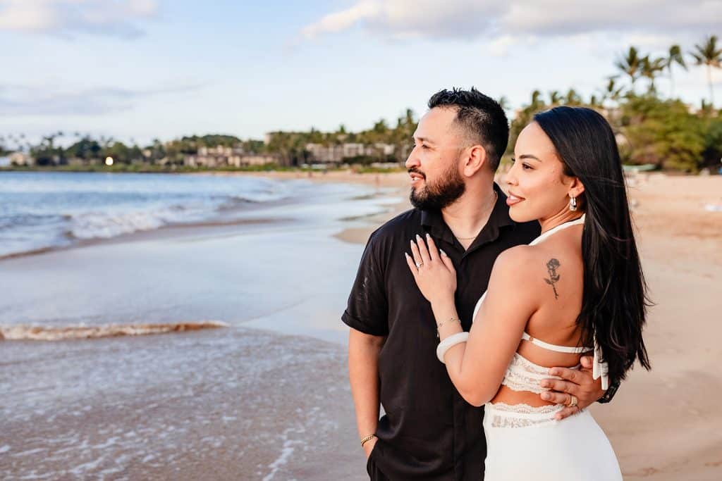 Brandon and Alexis standing at the shore and looking at the sunset. Brandon has his hand around her back and she is resting her hand on his chest.