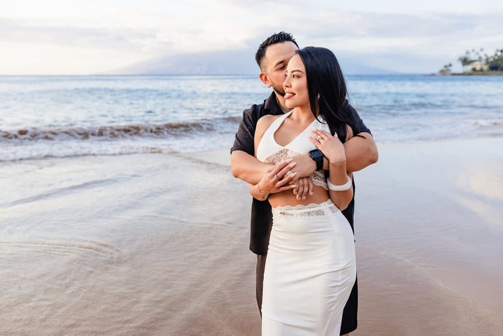 Brandon hugging Alexis from behind as she holds his arms around her and leans back into him. The ocean is behind them as he kisses her temple.