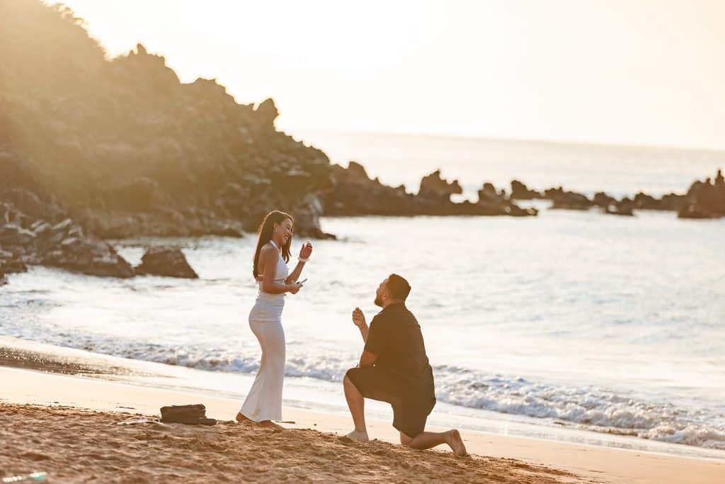 Brandon is down on one knee in the sand and holding a ring as Alexis is smiling down at him and bringing her hand up to her face.