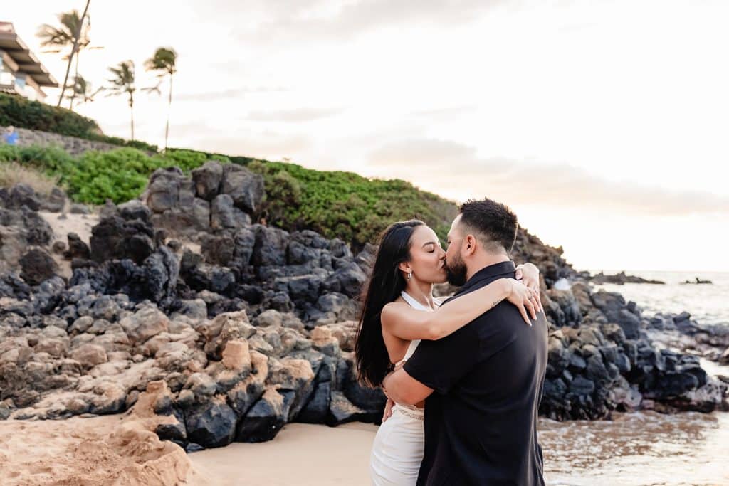 Brandon and Alexis sharing a kiss on the shoreline with the rocky cliff behind them.