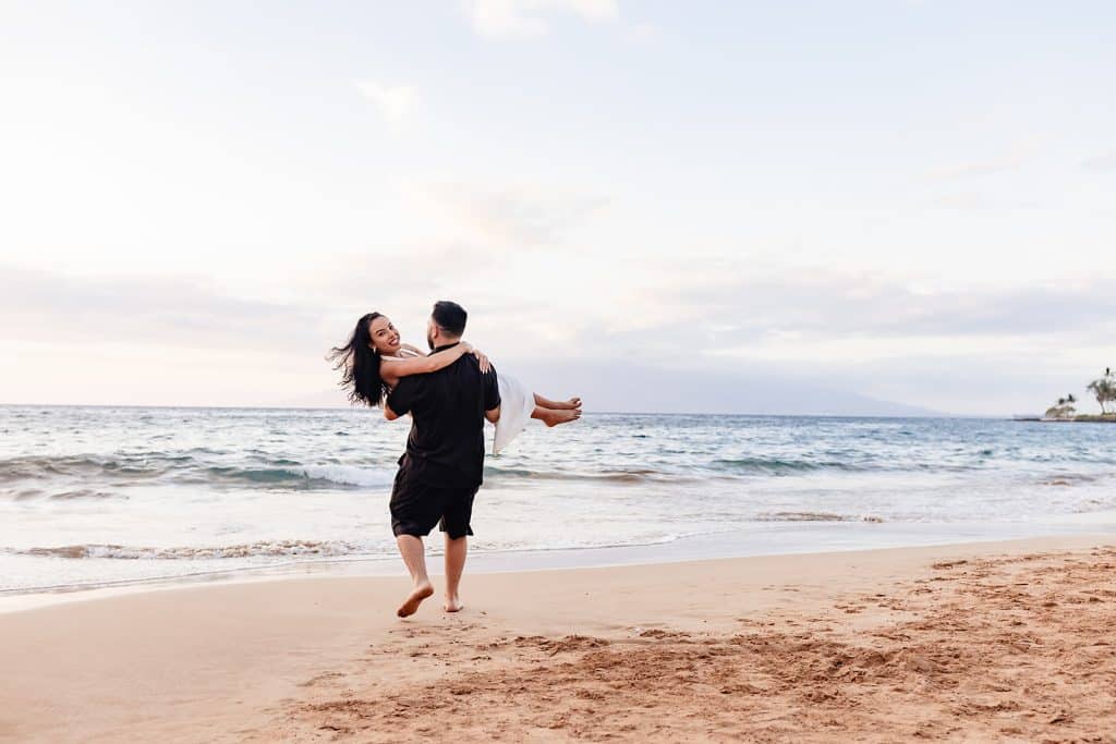 Brandon carrying Alexis and walking towards the water while she looks over his shoulder at the camera and smiles.