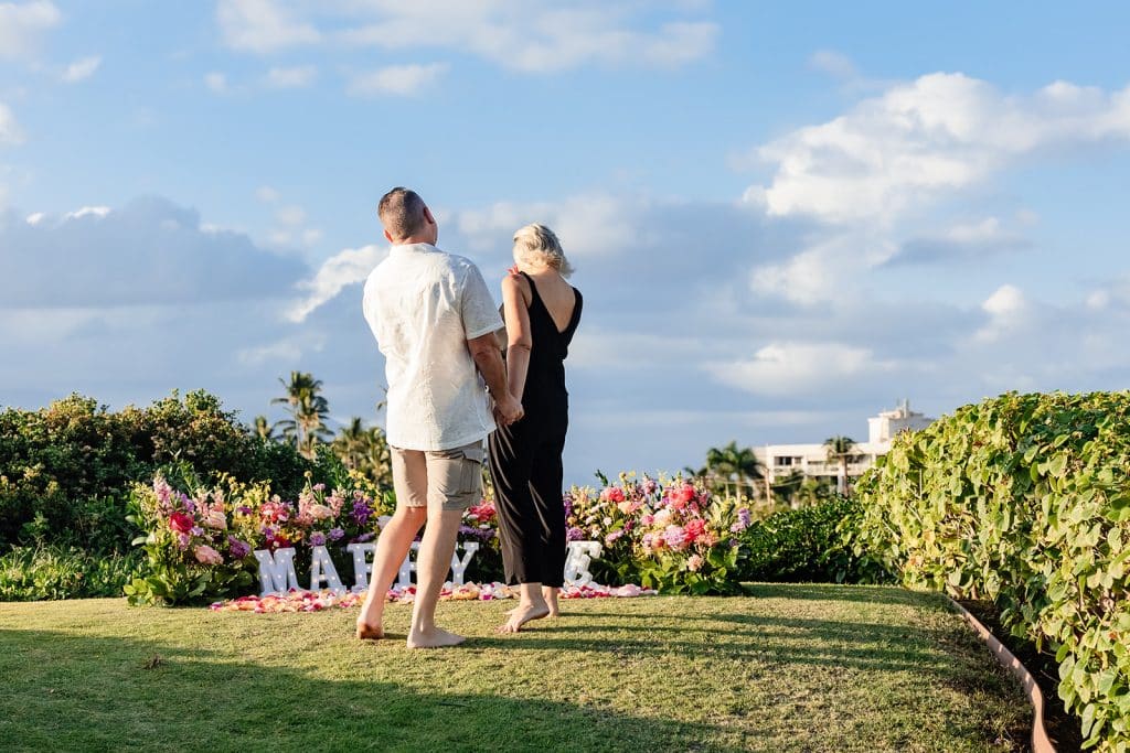 Andy and Joy walking up to the proposal set up. Joy is covering her mouth and looking at the flowers and marquee letters as Andy stands slightly behind her holding her hand.