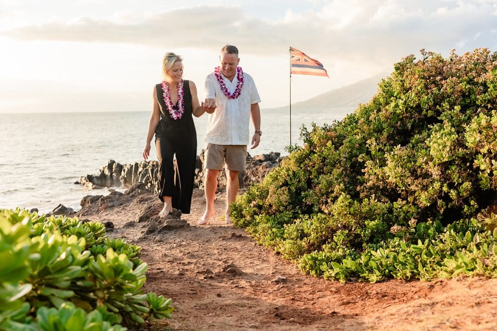 Joy and Andy holding hands and walking along the cliffside path. The ocean is behind them and greenery is all along the path on the right.