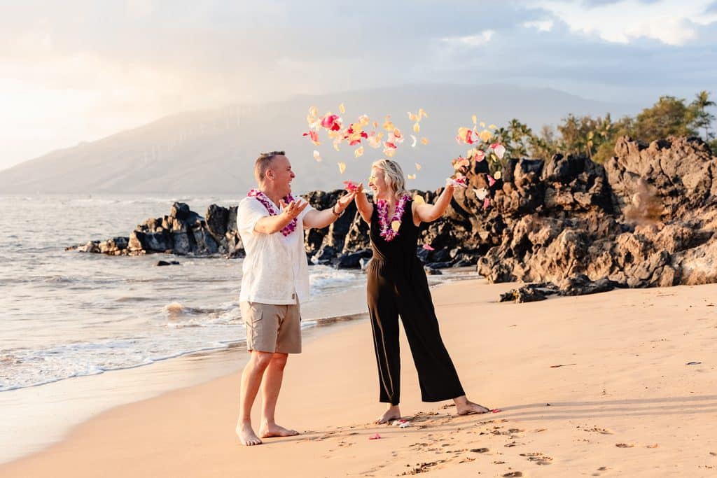 Joy and Andy standing on the beach and throwing flower petals into the air as they smile and laugh at each other.