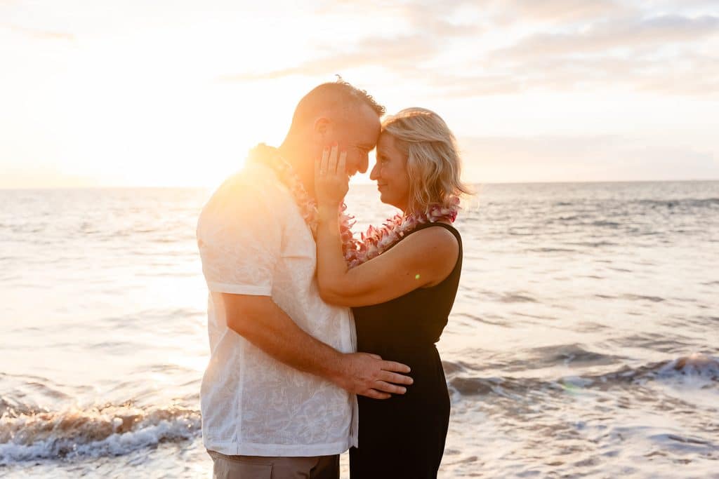 Joy holding Andy's face and smiling while he holds onto her waist. Their foreheads are touching and they are smiling at each other. The sun is setting over the ocean behind them leaving a golden glow.