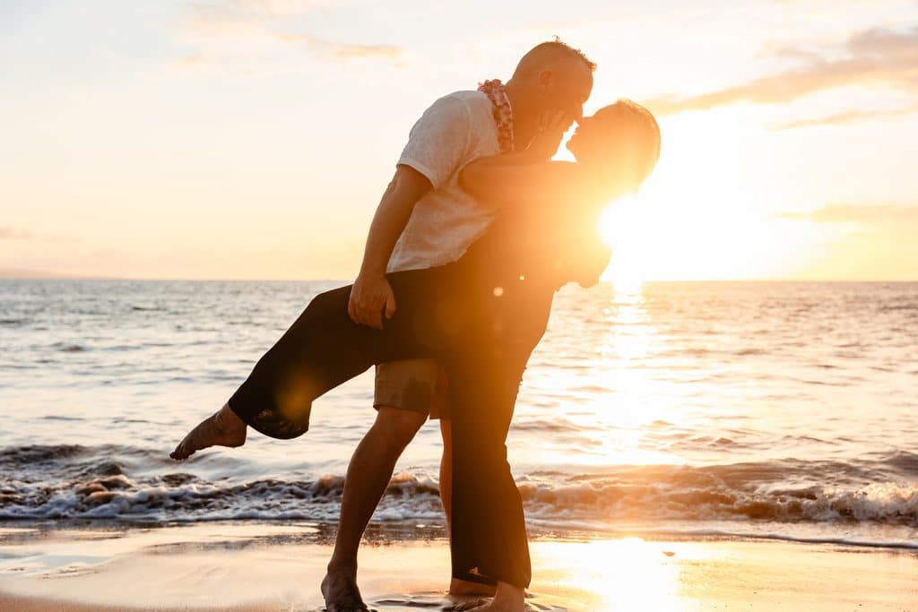 Andy dipping Joy for a kiss while holding one leg up. They're feet are in the sand and the sun is glowing behind them so it's almost just their silhouette. 