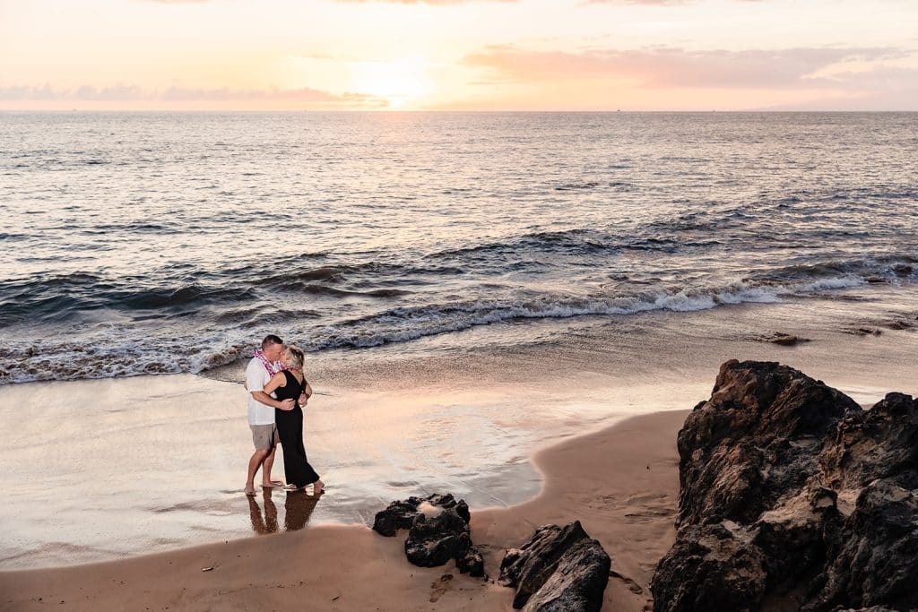 A wide photo from above of Andy and Joy kissing in the sand as the sun sets behind them.
