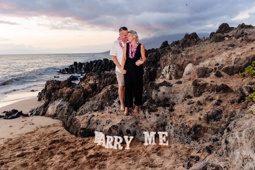 Andy holding Joy from behind and nuzzling into her neck as they stand on coastal rocks. The 'Marry Me' marquee letters displayed at the bottom of the rocks in the sand.