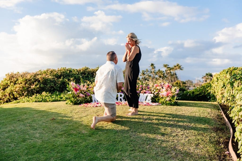 Joy turning around to see Andy on one knee. The flowers and 'Marry Me' letters are behind her as she holds her hands up to her face in shock.
