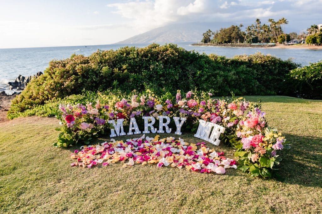 The proposal set up of colorful pink, white, and purple flowers sticking out of the ground. Marquee letters saying "Marry Me" are in front of the proposals and flower petals are in front of the marquee letters. The ocean and beach is behind the setup.