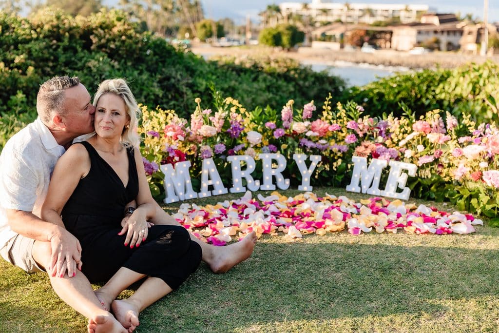 Andy and Joy sitting next to the proposal flowers and marquee letters. Joy is leaning into Andy who is sitting behind her. She's smiling at the camera while he kisses her cheek.