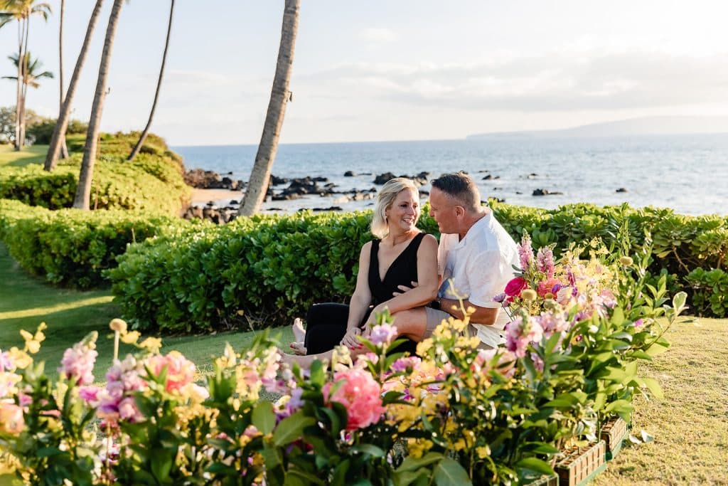 With the proposal flowers in front of Joy and Andy, they're looking at each other and smiling and talking while sitting on the ground next to the florals.