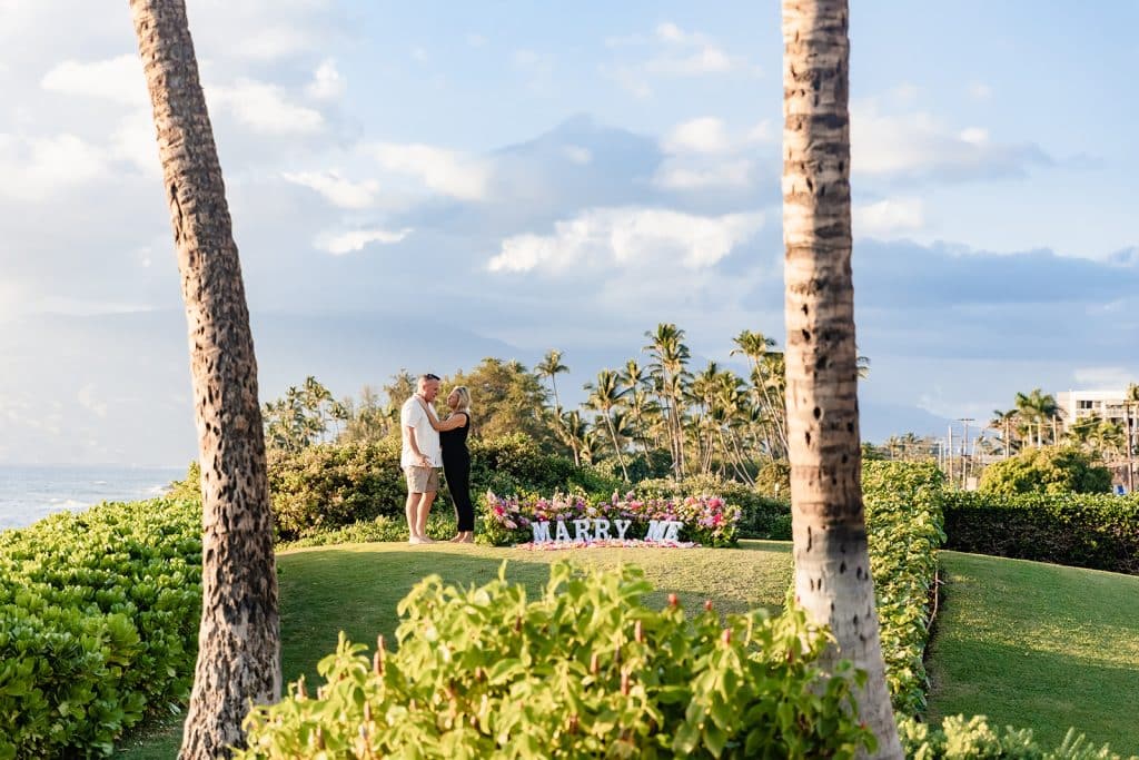 A wide photo of Andy and Joy embracing next to the proposal setup. They're in between two palm trees and the coast is behind them as the sun sets.