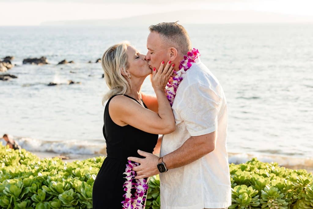 Joy giving Andy a kiss after putting his lei on. He holds her waist with a lei in his hand to place onto her neck. The ocean in the background is glistening with the sunset.