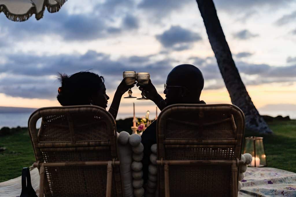 Ernest and Gloria's silhouettes sitting at the picnic in the picnic chairs and clinking their glasses to cheers as the sun fades behind the horizon at their sunset picnic proposal.