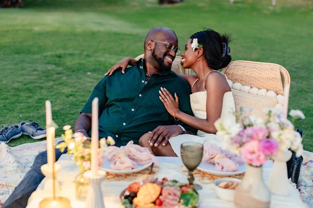 Gloria leaning into Ernest as they are sitting at their sunset picnic proposal setup and smiling at him while he smiles and rests his hand on her leg.