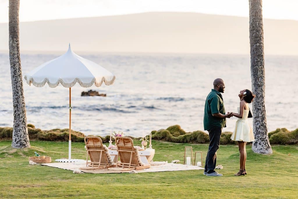 Ernest holding Gloria's hands next to the sunset picnic proposal setup and Gloria laughing at what he is saying.