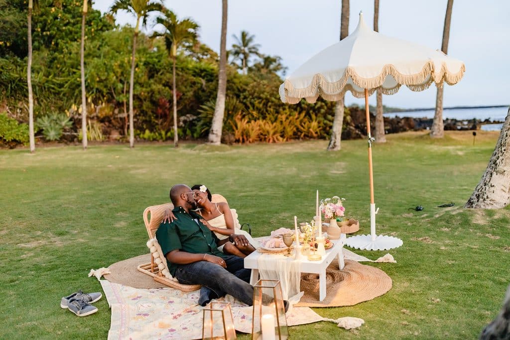 A far away photo of Ernest and Gloria sitting at the full sunset picnic proposal spread and leaning into each other smiling.