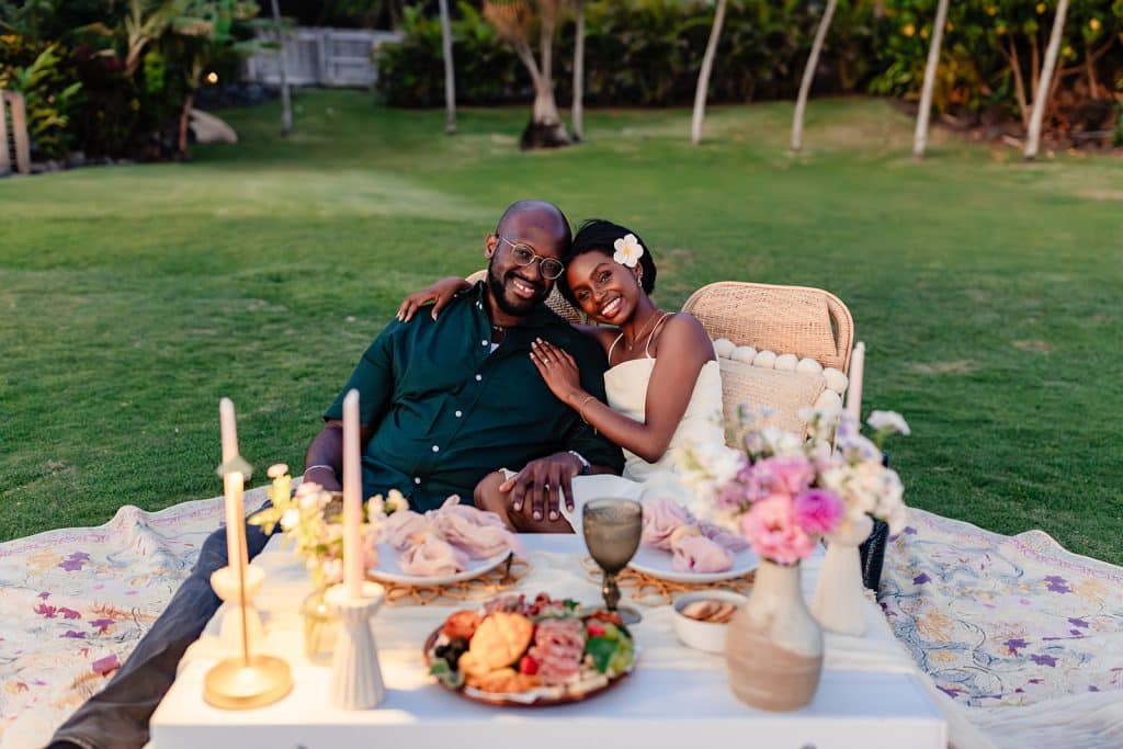 Ernest and Gloria sitting behind the picnic table and leaning into each other as the smile for the camera as their sunset picnic proposal comes to an end.