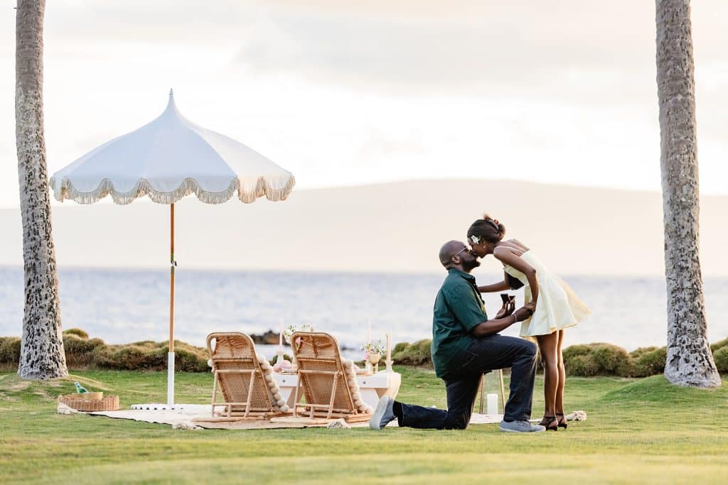 Ernest down on one knee and holding Gloria's hand with the ring box open in the opposite hand. Gloria is leaning down to give Ernest a kiss while he's proposing at his sunset picnic proposal.