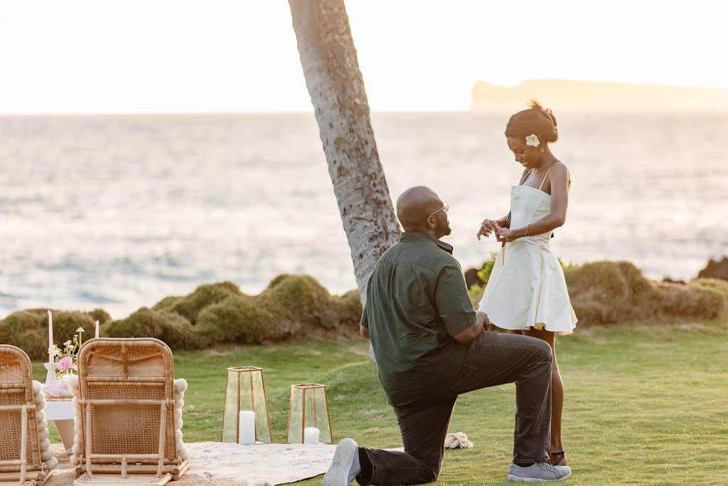 Ernest down on one knee in the grass next to the sunset picnic proposal setup as Gloria stares at the newly placed ring on her finger, smiling down at it.