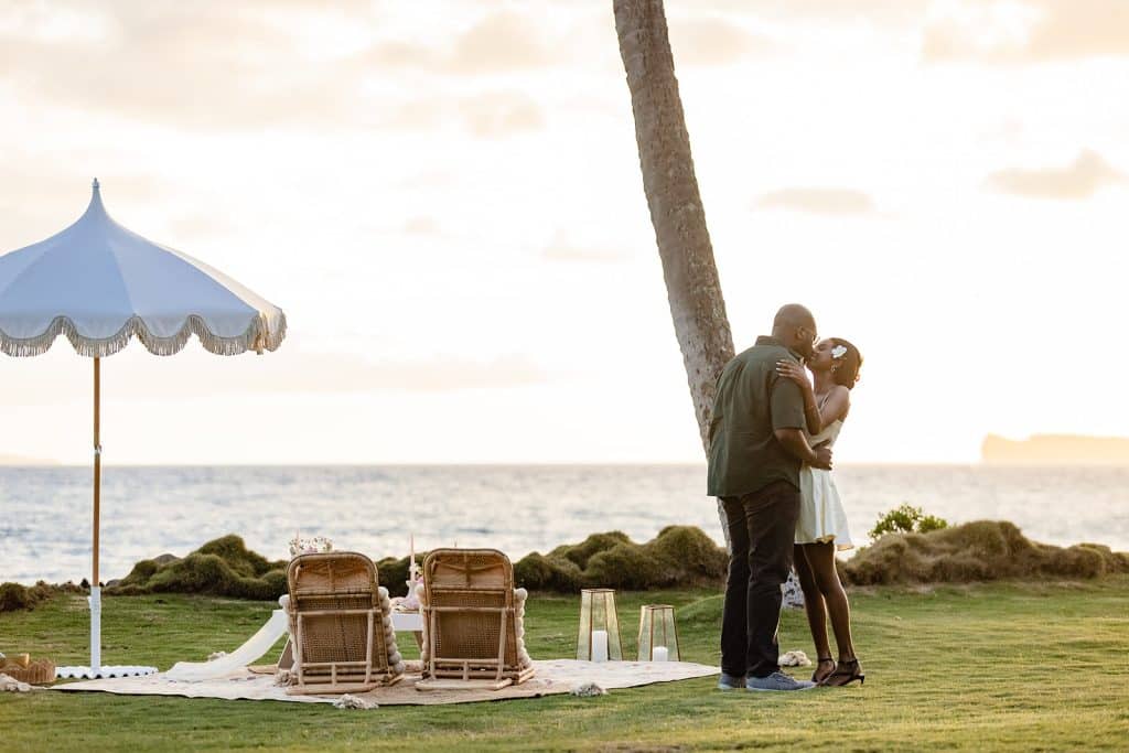Ernest and Gloria sharing a kiss after the proposal during his sunset picnic proposal with the beach in the background.