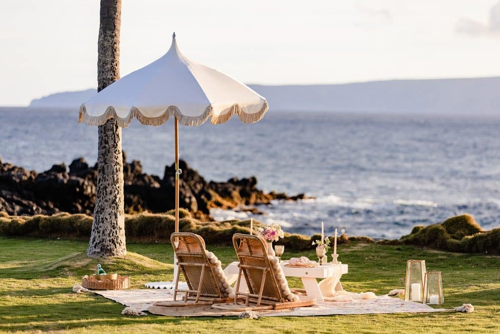 A sunset picnic proposal setup on the coast of Maui facing the ocean. Comfy floor chairs, a big umbrella, and a small table all decorated and shining in the sunset.